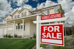 A two-story suburban home with a "Foreclosure For Sale" sign in the front yard, set against a partly cloudy blue sky.